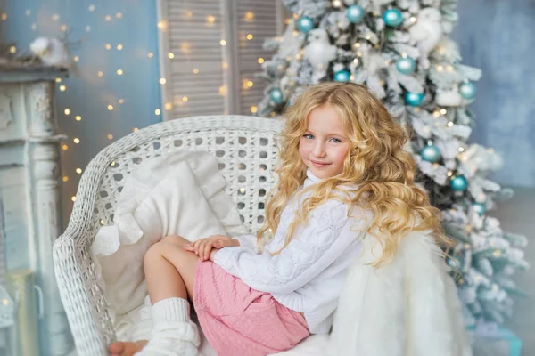 Pretty blonde little girl sits on a chair near a fireplace and Christmas tree — Stock Photo, Image