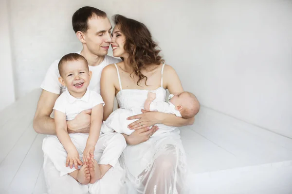 Retrato de una joven familia feliz vestida de blanco con un bebé y un niño en un estudio blanco —  Fotos de Stock