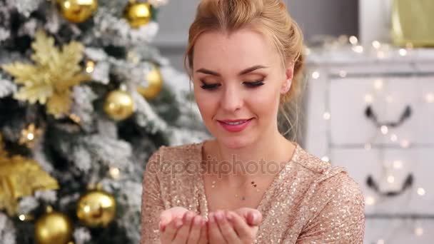 Beautiful blonde young woman blows a confetti from hands near Christmas tree — Stock Video