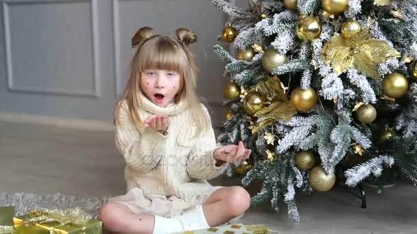 Emotional little girl sits under Christmas tree and blows a confetti from hands — Stock Video