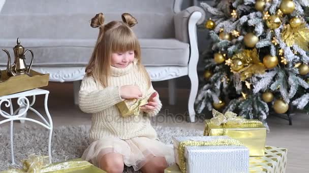 Emocional niña sostiene una caja de regalo en las manos, tratar de abrir cerca del árbol de Navidad en casa — Vídeo de stock