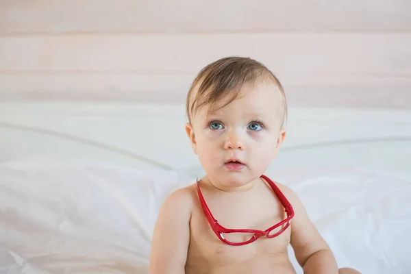 Pequeno bebê triste com uns óculos vermelhos em uma cama — Fotografia de Stock