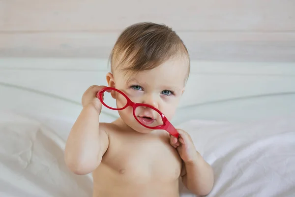 Retrato de un bebé caucásico jugando con unas gafas rojas en una cama — Foto de Stock