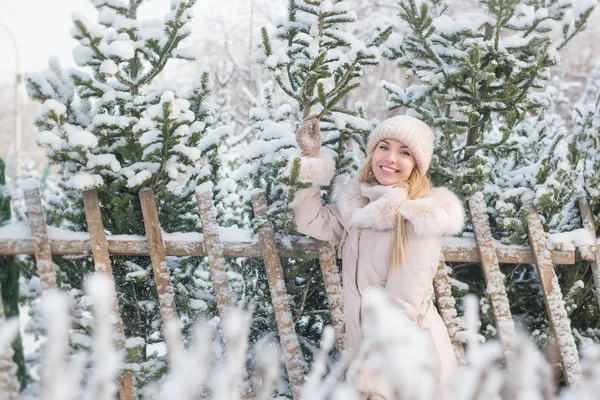 Портрет улыбающейся красивой девушки в бежевом пиджаке стоит возле елок в зимний день — стоковое фото
