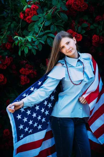 The young beautiful girl stands against the roses with the flag of the United States of America — Stock Photo, Image