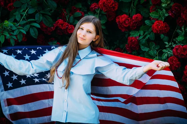 La giovane bella ragazza si erge contro le rose con la bandiera degli Stati Uniti d'America — Foto Stock