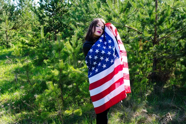 A young beautiful woman with a flag of the United States of America. Green forest.