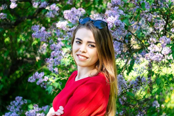 A menina na primavera vestida com uma camisola vermelha é em torno de um lilás azul . — Fotografia de Stock