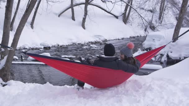 ユタ州の小さな川の横の雪に囲まれた冬の風景の中にハンモックで揺れるカップル — ストック動画