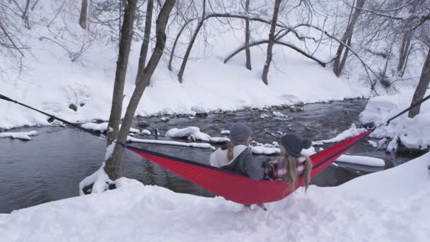 冬のために暖かい服装をしているので 雪の中で川の横にハンモックでリラックスしている2人の若い女性 — ストック動画