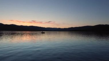 Alacakaranlıkta bir gölün üzerinden uçarken küçük bir balıkçı teknesinin yanından geçerken Montana 'da alacakaranlıkta bir kişi oltasını çekiyor..