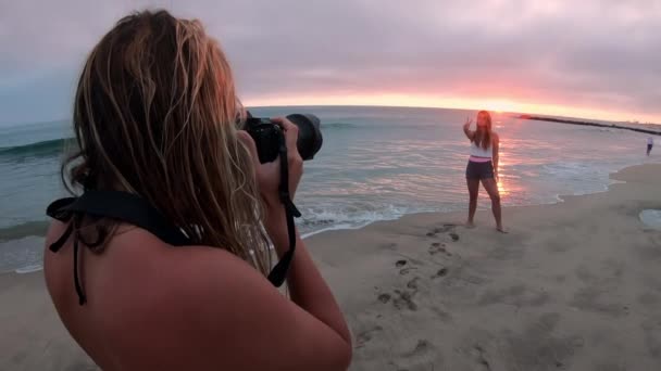 夕暮れ時にビーチで平和サインをしている若い女性と砂の中で彼女の写真を撮る女性 — ストック動画
