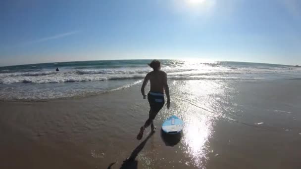 若い男がビーチで走り ビーチで海にそれに乗るボード上でジャンプします — ストック動画