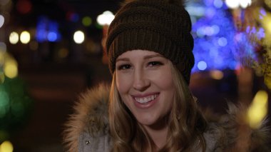 Genç bir kadın gece vakti Noel ışıklarının önünde gülümsüyor..