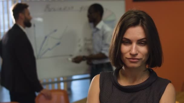 Joven empresaria sonriendo en una cámara, mientras sus colegas están haciendo su trabajo . — Vídeos de Stock