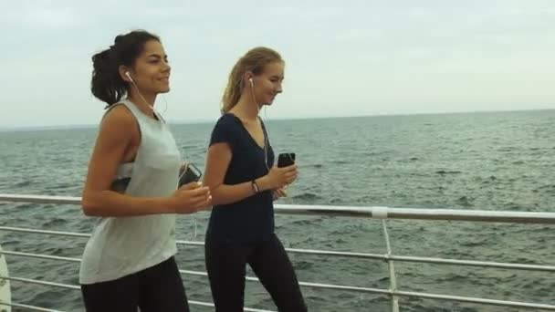Two athletic woman running outdoors in slow motion on promenade at sunset near ocean enjoying evening run — ストック動画