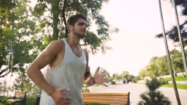 Grand jeune homme actif jogging sur le parc, tout en écoutant de la musique dans les écouteurs du smartphone lecteur mp3 . — Video