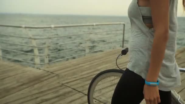 Schöne junge Frau mit dunkler Haarfarbe auf einem Fahrrad in der Nähe des Meeres auf der Holzpromenade. — Stockvideo