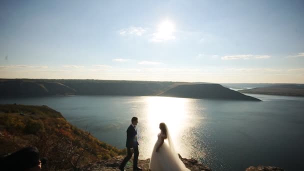 Junger hübscher Bräutigam im Anzug und die Braut im weißen eleganten Kleid am Ufer eines Bergsees. — Stockvideo