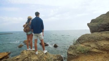 Genç bayan bir şapka ve küçük sırt çantası ve dalgalar, bir taş üstüne kalırken izlerken uzun boylu yakışıklı erkek tabaklanmış.