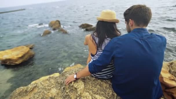 Dos jóvenes turistas felices sentados en las piedras, observando el mar en calma. Hombre abrazando a su mujer . — Vídeos de Stock