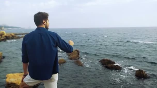 Jeune homme avec une coiffure drôle, rester sur le dessus d'une pierre et regarder la beauté de la mer . — Video