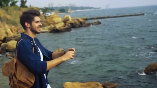 Jeune beau touriste heureux avec un sac à dos brun prenant des photos de belle mer calme bleu . — Video