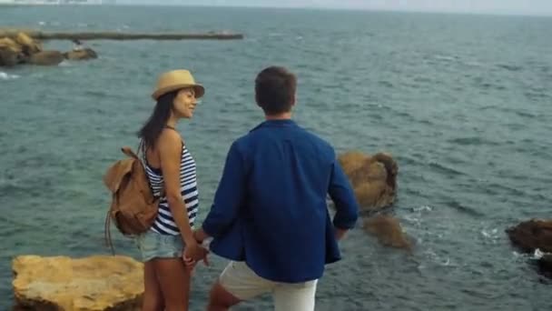 Mujer bonita sonriente sosteniendo una mano de su novio, permaneciendo en la piedra y mirando el mar . — Vídeos de Stock