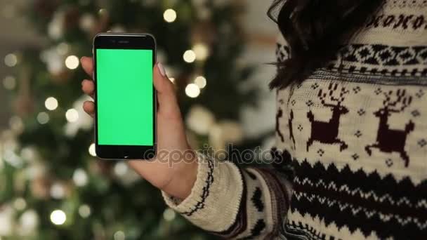 Крупный план женских рук, касающихся смартфона. Зеленый экран Chroma Key. Закрывай. Отслеживаю движение. на фоне размытого рождественского оформления — стоковое видео