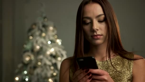 Süße lächelnde Dame in Goldkleid, posiert mit Smartphone an den Händen in der Nähe des Weihnachtsbaums vor der Kamera. — Stockvideo