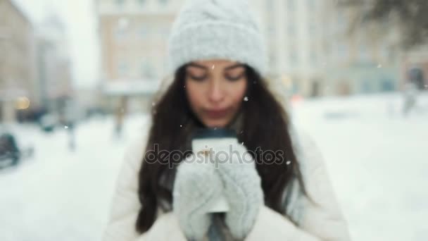 Geluk, wintervakantie, Kerstmis, dranken en mensen concept - jonge vrouw in witte warme kleren met glimlachen en drinken koffie weg te nemen over besneeuwde stad achtergrond — Stockvideo