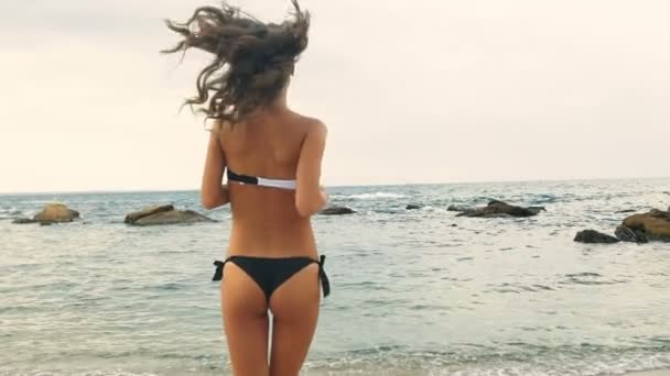 Νέοι σέξι κυρία σε μαύρο και άσπρο μαγιό που τρέχει στο νερό σε μια παραλία. — Αρχείο Βίντεο