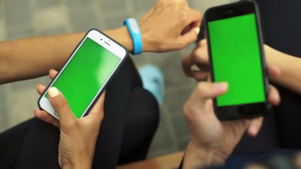 Dos deportistas sentadas en un banco, sosteniendo dos teléfonos inteligentes en las manos. Llave de croma de pantalla verde. De cerca. . — Vídeos de Stock