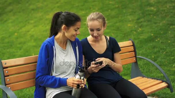 Two young girls sitting on a bench, one girl holding smartphone, other one vacuum bottle. — Stock Video