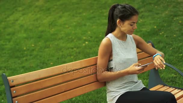 Jovem bela mulher morena sentada em um banco e usando rastreador de atividades em um parque . — Vídeo de Stock