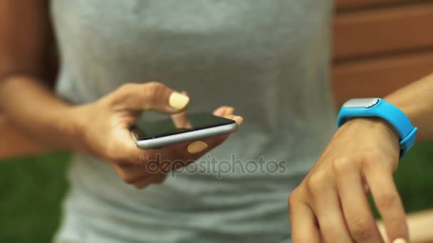 Jovencita con bonitas uñas amarillas sentada en un banco con brazalete deportivo y teléfono inteligente . — Vídeo de stock