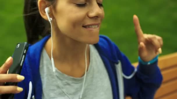 听音乐对她的耳机，坐在一条长凳上的年轻女士的特写. — 图库视频影像