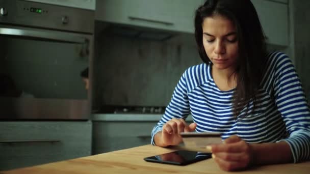 Молода жінка сидить на кухні, тримає карту в руках і робить покупки онлайн . — стокове відео