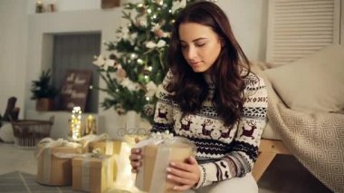 Genç, mutlu, şaşkın kadın süslü Noel ağacının yanındaki hediye kutusunu açıyor.