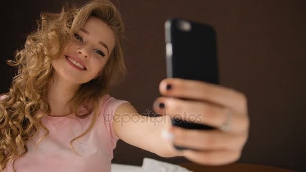 Jeune femme blonde prenant des selfies, assise sur le lit en pyjama rose . — Video