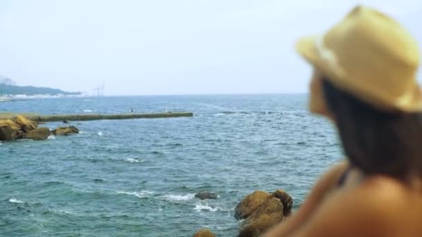 Jovem mulher feliz sentada na pedra e olhando para as ondas no mar . — Vídeo de Stock