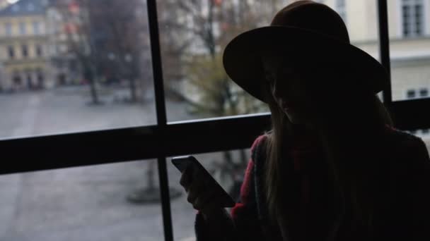 Silhouet van de vrouw gebruik mobiele telefoon in café van de stad bij zonsondergang op onscherpe achtergrond van architectuur en weg. 4k — Stockvideo