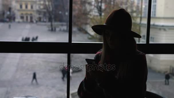 Silhouette of woman use mobile phone in city cafe at sunset on blurred architecture and road background. 4k — Stock Video