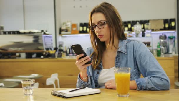 Молода красива жінка друкує на смартфоні і п'є сік в кафе — стокове відео