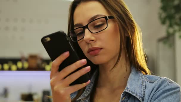 Mooie jonge vrouw in glazen met smartphone, SMS berichten in café — Stockvideo