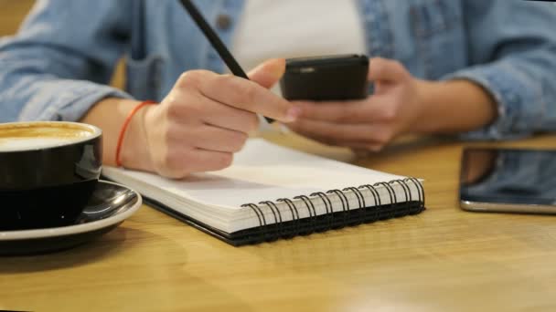 Закройте руки женщине, держащей смартфон и пишущей в блокноте в кафе — стоковое видео