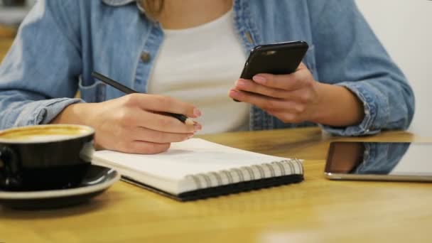 Крупним планом жіночі руки тримають смартфон і пишуть в блокноті в кафе з дерев'яними меблями — стокове відео