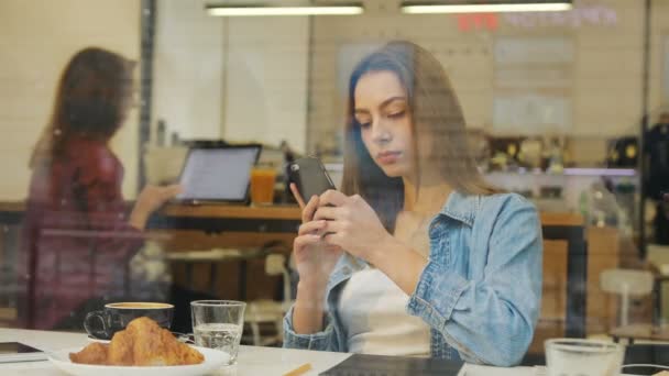 Όμορφη νεαρή γυναίκα, χρησιμοποιώντας το smartphone στο καφενείο, δείτε μέσα από το παράθυρο. — Αρχείο Βίντεο