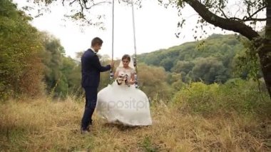 Damat güzel gelini sallanan beyaz gelinlik içinde çiçek buketi yaz ormandaki salıncak üzerinde ile park