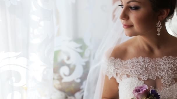 Beautiful bride in luxury wedding dress looking through the window before wedding ceremony. Woman holding flowers — Stock Video